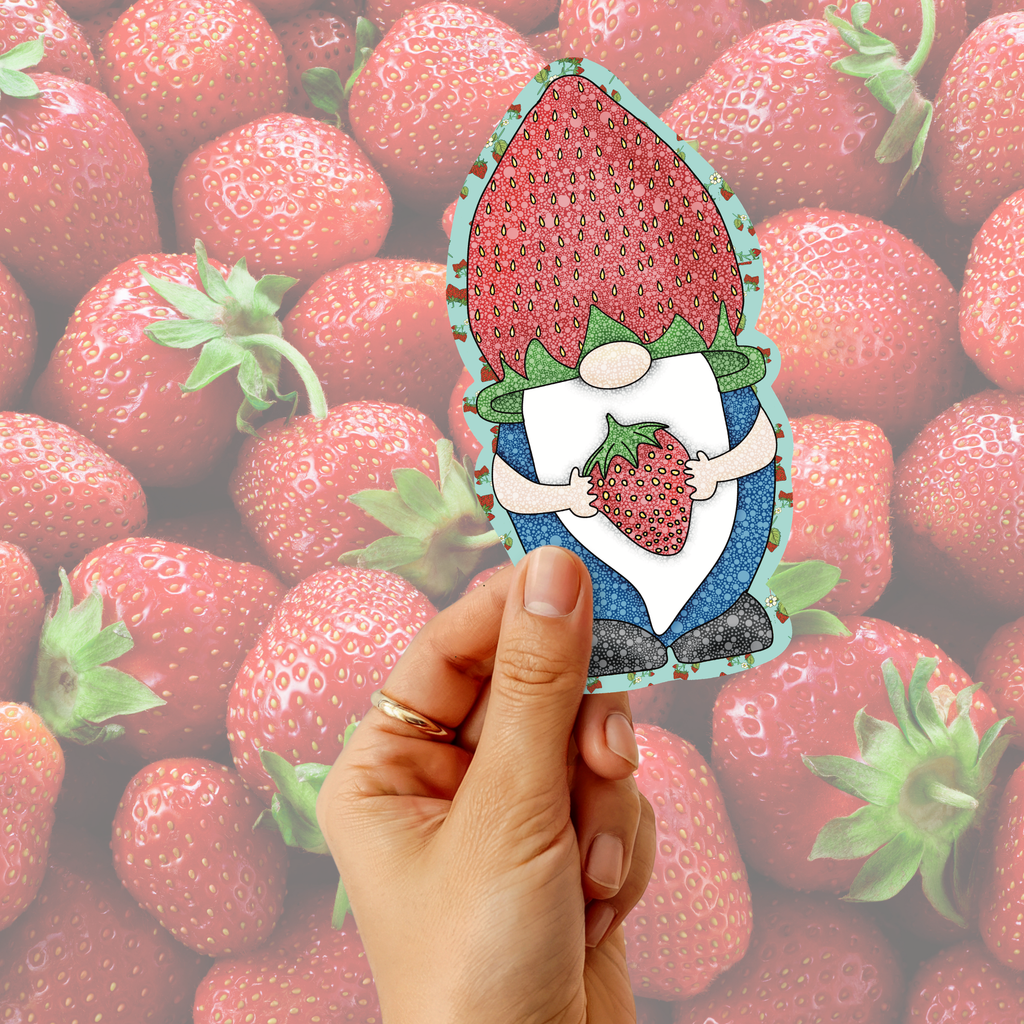 A hand holds a die-cut sticker or card featuring a gnome character with a large strawberry on its head and holding a strawberry in its hands, set against a background of fresh strawberries.