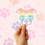 A hand holds a die-cut sticker shaped like a dog head with an ornate floral line-art design in a rainbow gradient. The background is a soft pink with pastel paw prints.