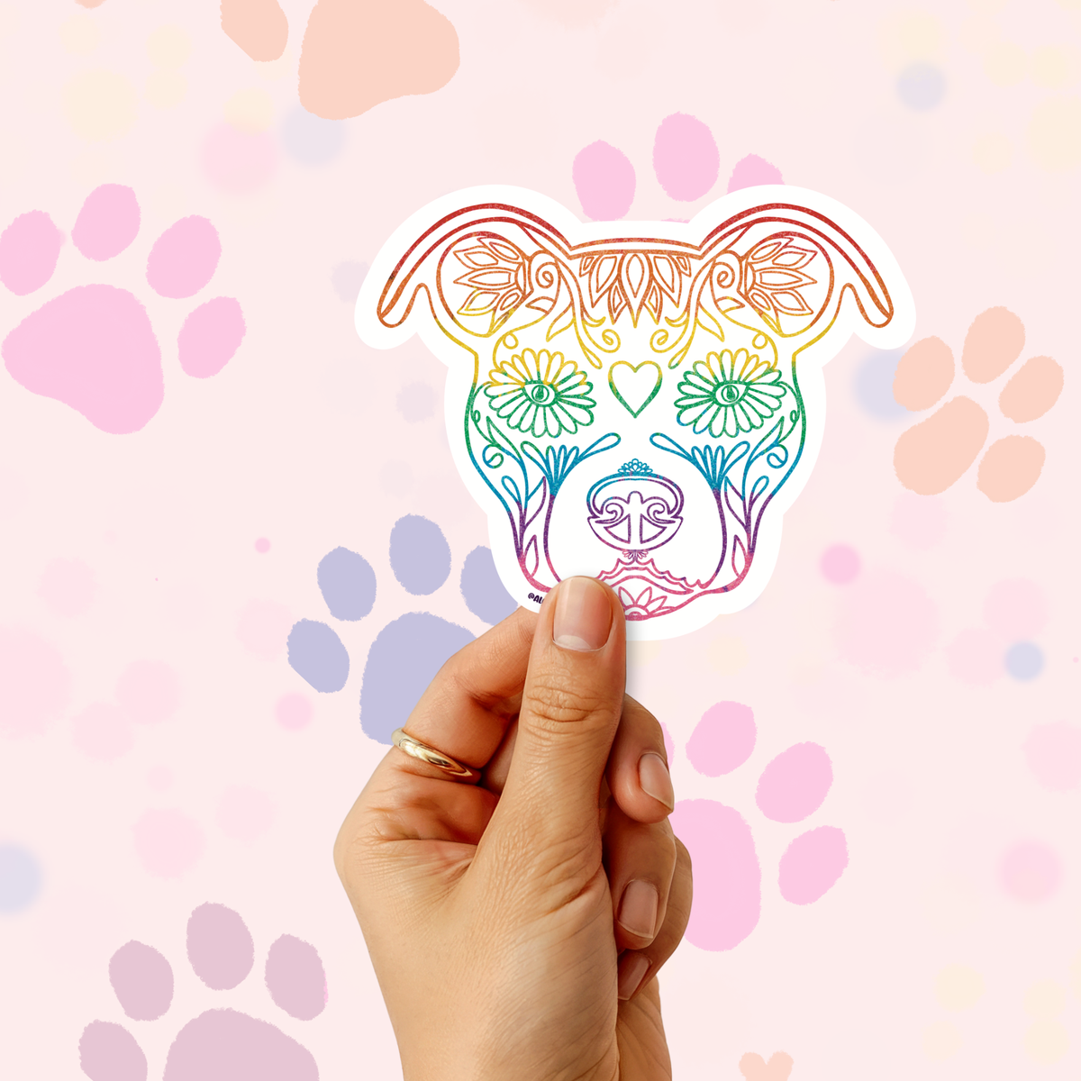 A hand holds a die-cut sticker shaped like a dog head with an ornate floral line-art design in a rainbow gradient. The background is a soft pink with pastel paw prints.