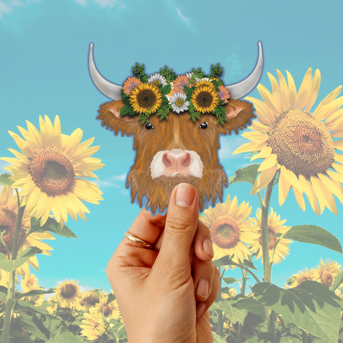 A hand holds a die-cut sticker or decal shaped like a brown Highland cow with white horns and a flower crown, set against a sunflower field background with a blue sky.