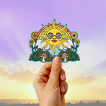 A hand holds a colorful die-cut sticker featuring a smiling sun wearing sunglasses, surrounded by flowers, mushrooms, and green leaves, against a sunset sky background.