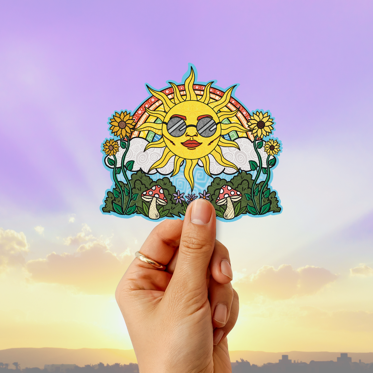A hand holds a colorful die-cut sticker featuring a smiling sun wearing sunglasses, surrounded by flowers, mushrooms, and green leaves, against a sunset sky background.