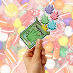 A hand holds a green die-cut sticker featuring a cannabis leaf and the words 'Sweet treats'  with four colorful flower-shaped candies placed on top, against a background of assorted wrapped lollipops and candies.