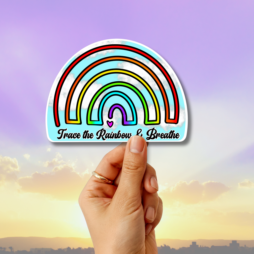 A hand holds a die-cut sticker featuring a colorful rainbow design with the text 'Trace the Rainbow Breathe' against a sunset sky background.