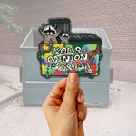 A hand holds a die-cut sticker in front of a gray dumpster in an outdoor alley. The sticker features two raccoons above a colorful graffiti-style background with the text 'YOUR OPINION IS TRASH'.