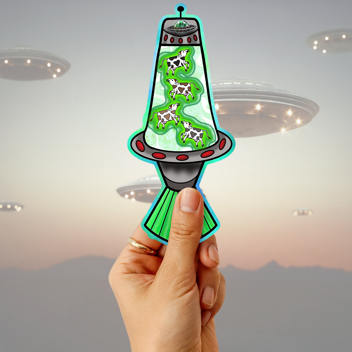 A hand holds a die-cut sticker shaped like a retro flying saucer with green liquid and four black-and-white cows inside, set against a blurred sunset sky with UFOs.