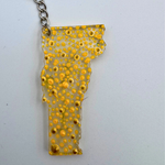 A clear acrylic pendant shaped like the state of Vermont, filled with small yellow and gold bubble-like dots, hanging from a silver chain against a plain light background.