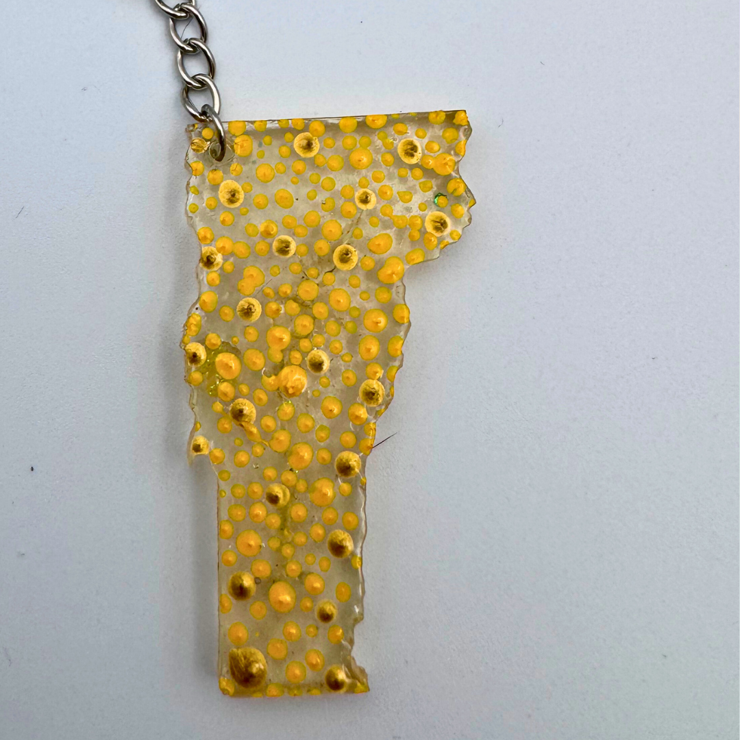 A clear acrylic pendant shaped like the state of Vermont, filled with small yellow and gold bubble-like dots, hanging from a silver chain against a plain light background.