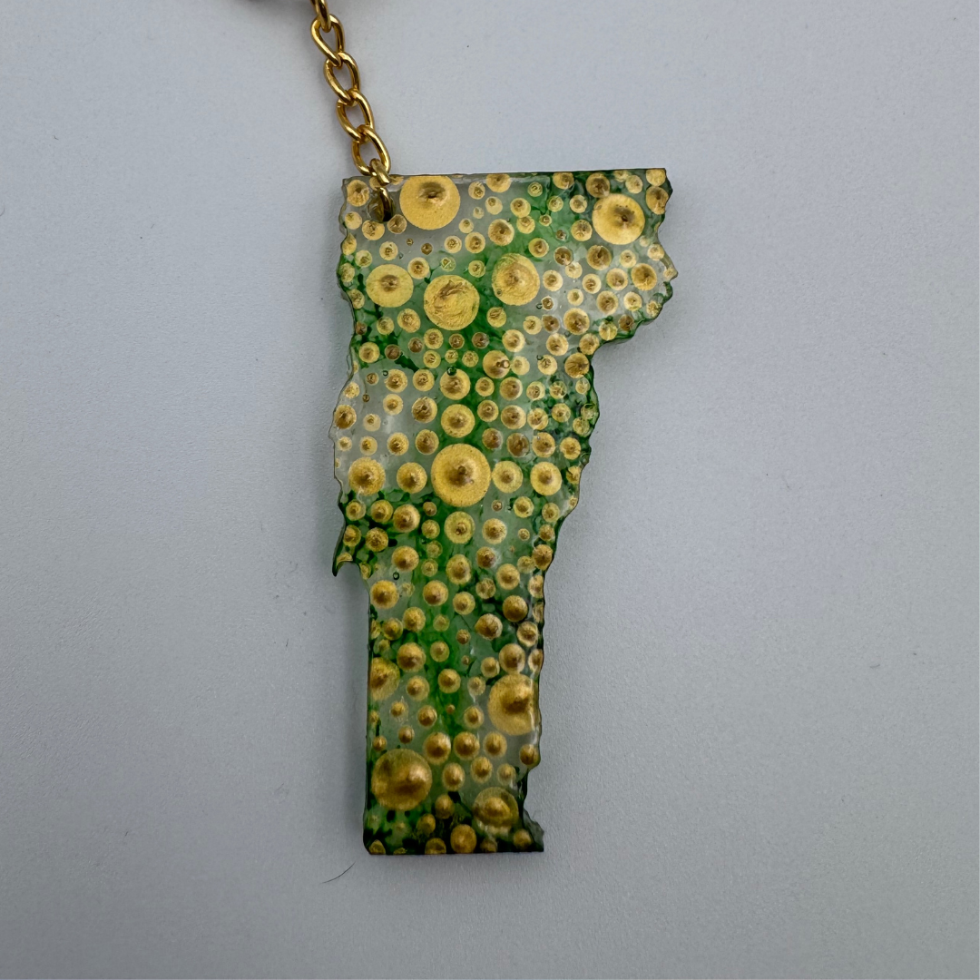 A Vermont-shaped keychain featuring a green and gold spotted pattern, suspended from a gold chain against a light gray background.