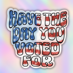 A die-cut sticker with the text 'HAVE THE DAY YOU VOTED FOR' in bold bubble letters filled with an American flag pattern of blue, red, and white, outlined in black, shown on a pastel rainbow gradient background.